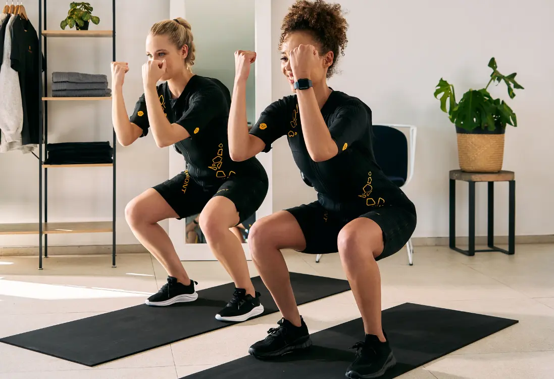 Snelle resultaten met EMS training in Hilversum Twee vrouwen in EMS-pakken doen squats en gebruiken EMS-training in Hilversum om in korte tijd snelle resultaten te boeken.